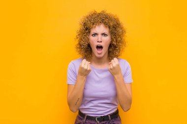 Genç afro kadın sinirli sinirli bakışlar ve sıkı yumruklarla bağırıyor, turuncu duvara karşı öfkeli hissediyor.