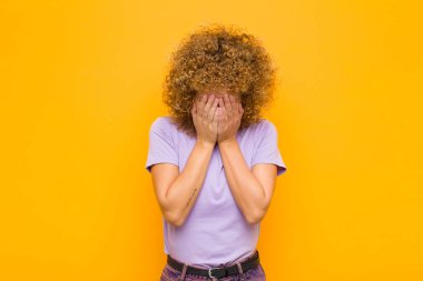 Genç afro kadın üzgün, hayal kırıklığına uğramış, gergin ve depresif, yüzünü iki eliyle kapatmış, turuncu duvara karşı ağlıyor.