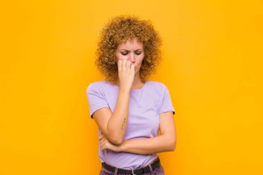 Genç afro kadın ciddi, düşünceli ve endişeli, elleri turuncu duvara yaslanmış bir şekilde yan bakıyor.