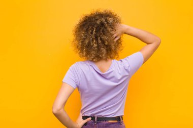 Genç afro kadın düşünüyor ya da şüphe ediyor, kafasını kaşıyor, şaşkın ve şaşkın hissediyor, turuncu duvarın arkasında ya da arkasında