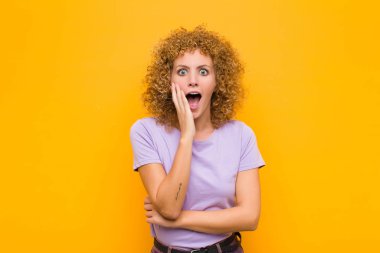 Genç afro kadın şok ve inançsızlık içinde, elleri yanak ve kolları çaprazlanmış, sersemlemiş ve turuncu duvara karşı hayrete düşmüş.