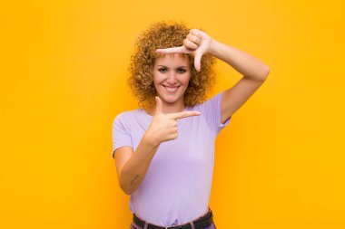 Genç afro kadın mutlu, arkadaş canlısı ve pozitif hissediyor, gülümsüyor ve portre veya fotoğraf çerçevesi yapıyor. Elleri turuncu duvara dayalı.