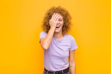 Genç Afro kadın gülüyor ve alnına şaplak atıyor. Unutmuşum yoksa turuncu duvara karşı yaptığım aptalca bir hataydı.