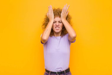 Genç afro kadın stresli ve endişeli, depresif ve hayal kırıklığına uğramış hissediyor, iki elini de turuncu duvara karşı kaldırıyor.