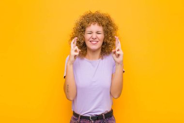 Genç afro kadın gülümsüyor ve endişeyle iki parmağını çapraz yapıyor. Endişeli ve turuncu duvara karşı iyi şanslar diliyor.