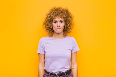 Genç afro kadın şaşkın ve kafası karışmış görünüyor. Gergin bir hareketle dudağını ısırıyor. Turuncu duvardaki sorunun cevabını bilmiyor.