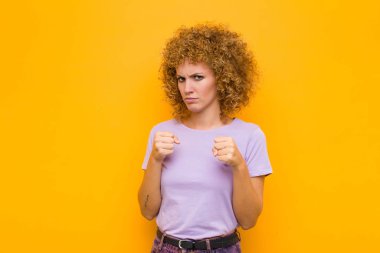 Genç afro kadın kendinden emin, sinirli, güçlü ve agresif görünüyor. Yumrukları turuncu duvara karşı boks pozisyonunda dövüşmeye hazır.