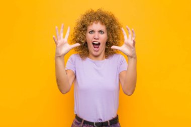 Genç afro kadın ellerini havaya kaldırarak bağırıyor. Öfkeli, sinirli, stresli ve turuncu duvara karşı üzgün.