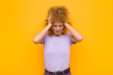 Genç afro kadın stresli ve hayal kırıklığına uğramış hissediyor, el ele tutuşuyor, yorgun, mutsuz ve turuncu duvarda migreni var.