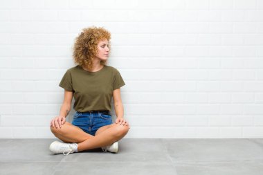 Genç afro kadın düşünüyor ya da şüphe ediyor, kaşınıyor, kafası karışık ve şaşkın hissediyor, arka ya da arka penceresi yerde oturuyor.