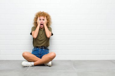 Genç afro kadın hoş olmayan bir şekilde şok olmuş, korkmuş ya da endişeli görünüyor, ağzı sonuna kadar açık ve elleri yerde oturan iki kulağını da kapatıyor.