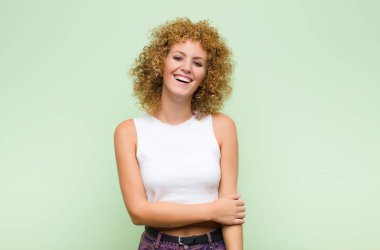Genç afro kadın utangaç ve neşeli bir şekilde gülüyor, yeşil duvara karşı arkadaş canlısı ve olumlu ama güvensiz bir tutumla