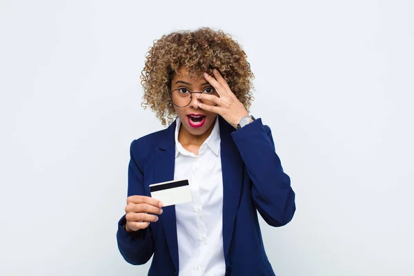 アフリカ系アメリカ人の若い女性はショックを受け恐怖に怯え顔を手で覆いクレジットカードで指の間を覗き — ストック写真