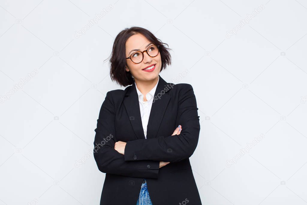 young businesswoman feeling happy, proud and hopeful, wondering or thinking, looking up to copy space with crossed arms against white wall