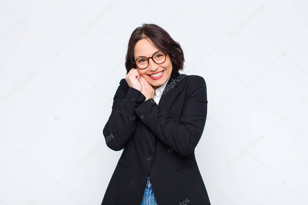 young businesswoman feeling in love and looking cute, adorable and happy, smiling romantically with hands next to face against white wall