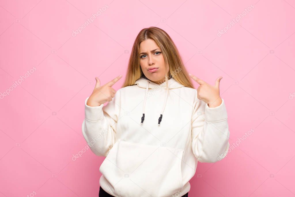 young blonde woman with a bad attitude looking proud and aggressive, pointing upwards or making fun sign with hands against flat wall