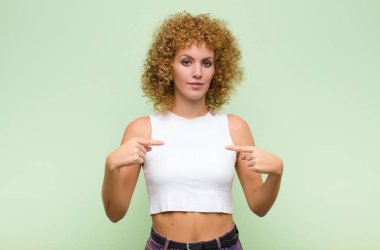 Genç afro kadın gururlu, pozitif ve rahat görünüyor. İki elini de yeşil duvara dayamış göğsünü gösteriyor.