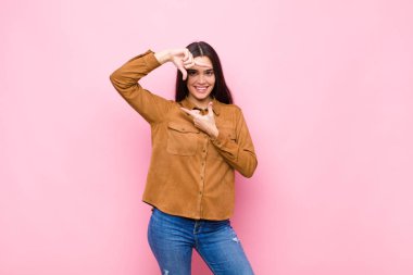 Genç, güzel bir kadın mutlu, arkadaş canlısı ve pozitif hissediyor, gülümsüyor ve elleri pembe duvara dayalı bir portre veya fotoğraf çerçevesi yapıyor.