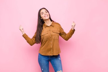 Genç ve güzel bir kadın son derece mutlu ve şaşırmış görünüyor, başarıyı kutluyor, bağırıyor ve pembe duvara zıplıyor.