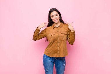 Genç, güzel bir kadın, kendine güvenerek gülümseyerek pembe duvara karşı pozitif, rahat ve tatmin edici bir tavır sergiliyor.