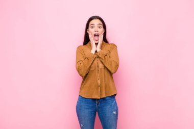 Genç ve güzel bir kadın şok olmuş ve korkmuş hissediyor. Ağzı açık ve elleri pembe duvarda.