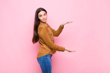 Genç ve güzel bir kadın, iki eli de kenarda olan bir objeyi tutuyor. Fotokopi alanı, pembe duvara karşı bir objeyi gösteriyor, sunuyor ya da reklamını yapıyor.