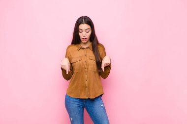 İki eliyle aşağıya bakan ağzı açık genç, güzel bir kadın. Pembe duvara karşı şaşkın, şaşkın ve şaşırmış görünüyor.