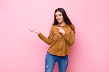 Genç ve güzel bir kadın neşeyle gülümsüyor ve kenardaki fotokopi alanını işaret ediyor, pembe duvara karşı bir nesneyi gösteriyor ya da reklamını yapıyor.