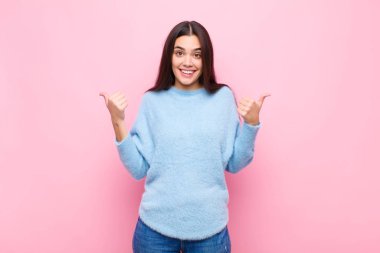 Genç, güzel bir kadın neşeyle gülümsüyor ve mutlu görünüyor. Kaygısız ve pozitif hissediyor. İki başparmağı da pembe duvarda.