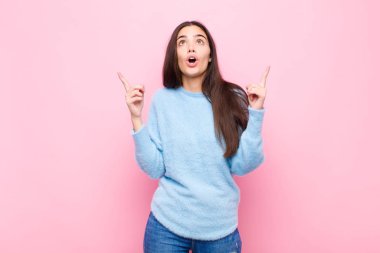 Genç, güzel bir kadın şaşırmış, şaşkın ve ağzı açık görünüyor, iki eliyle yukarıya bakıyor pembe duvara uzayı kopyalamak için.