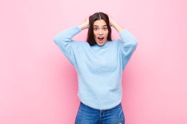 Genç ve güzel bir kadın heyecanlı ve şaşırmış görünüyor, ağzı açık, iki eli başının üstünde, pembe duvara karşı şanslı bir kazanan gibi hissediyor