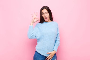 Genç, güzel bir kadın kendini başarılı ve tatmin olmuş hissediyor. Ağzı açık gülümsüyor. Elleri pembe duvara yaslanmış şekilde işaret veriyor.