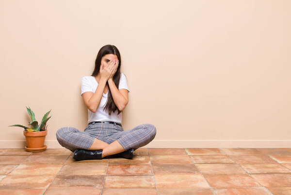 young pretty woman covering face with hands, peeking between fingers with surprised expression and looking to the side sitting on a floor