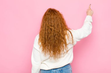 Genç kızıl saçlı kadın dikiliyor ve fotokopi alanındaki objeyi işaret ediyor. Pembe duvara karşı dikiz manzaralı.