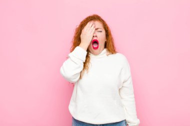 Genç kızıl saçlı kadın uykulu görünüyor, sıkılmış ve esniyor, baş ağrısı ve yüzünün yarısı pembe duvarla kaplı.