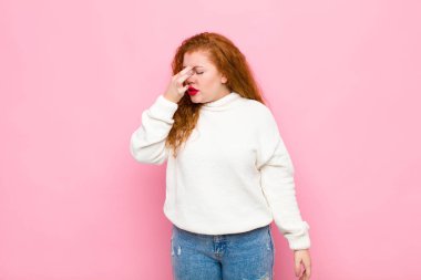 Genç kızıl saçlı kadın stresli, mutsuz ve hayal kırıklığına uğramış, alnına dokunuyor ve pembe duvara karşı şiddetli baş ağrısı çekiyor.