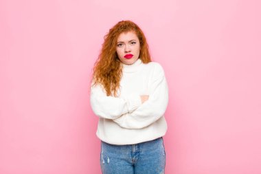 Genç kızıl saçlı kadın hayal kırıklığına uğramış ve hayal kırıklığına uğramış, ciddi görünüyor, kızgın ve pembe duvara karşı çapraz kollara sahip