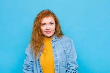 Genç kızıl saçlı kadın gururlu, kendinden emin, havalı, küstah ve küstah görünüyor, gülümsüyor, mavi duvara karşı kendini başarılı hissediyor.