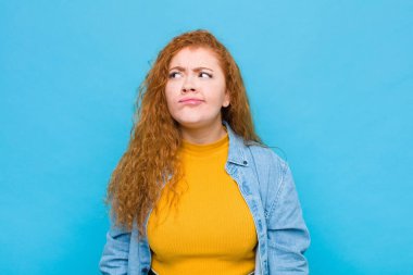 Genç kızıl saçlı kadın şaşkın ve şaşkın görünüyor. Bir sorunu çözmeye çalışıyor ya da mavi duvara karşı düşünüyor.