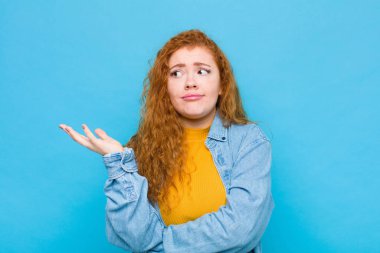 Genç kızıl saçlı kadın, kafası karışık ve bilgisiz hissediyor, şüpheli bir açıklamayı merak ediyor ya da mavi duvara karşı bir şey düşünüyor.