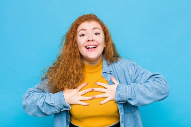 Genç kızıl saçlı kadın mutlu, şaşırmış, gururlu ve heyecanlı görünüyor, mavi duvara karşı kendini işaret ediyor.