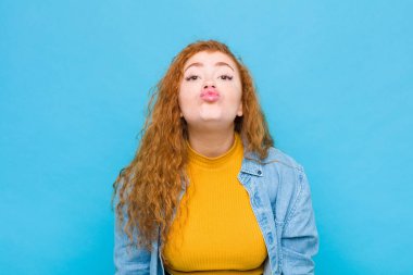 Genç kızıl saçlı kadın dudaklarını birbirine bastırıyor, tatlı, eğlenceli, mutlu, sevimli bir yüz ifadesiyle, mavi duvara bir öpücük yolluyor.