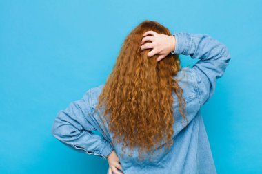 Genç kızıl saçlı kadın düşünüyor ya da şüphe ediyor, kafasını kaşıyor, şaşkın ve şaşkın hissediyor, mavi duvarın arkasında ya da arkasında.