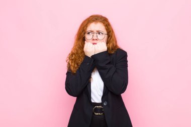 Genç kızıl saçlı kadın endişeli, stresli ve korkmuş görünüyor, tırnaklarını yiyor ve pembe duvara karşı yanal kopyalama alanı arıyor.