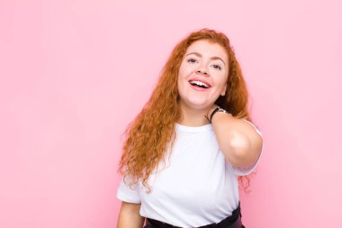 Genç kızıl saçlı kadın neşeli ve kendinden emin bir şekilde gülüyor pembe duvarın karşısında mutlu, samimi bir gülümseme ile.