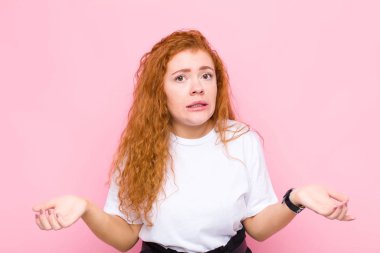 Genç kızıl saçlı kadın kendini cahil ve kafası karışık hissediyor. Hangi seçeneği seçeceğinden emin değil. Pembe duvara karşı merak içinde.