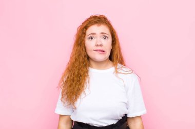 Genç kızıl saçlı kadın şaşkın ve şaşkın görünüyor, gergin bir hareketle dudaklarını ısırıyor, pembe duvara karşı sorunun cevabını bilmiyor.