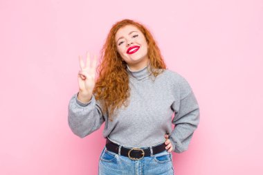 Genç kızıl saçlı kadın gülümsüyor ve arkadaş canlısı görünüyor. Üç ya da üç numarayı gösteriyor. Elleri önde, pembe duvara doğru sayıyor.