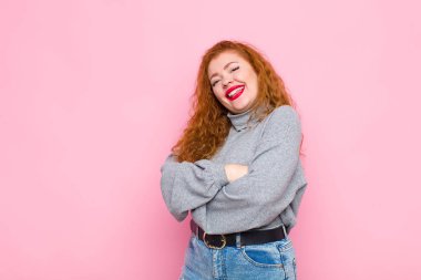 Genç kızıl saçlı kadın kollarını kavuşturup mutlu bir şekilde gülüyor. Pembe duvarın karşısında rahat, pozitif ve tatmin olmuş bir poz veriyor.