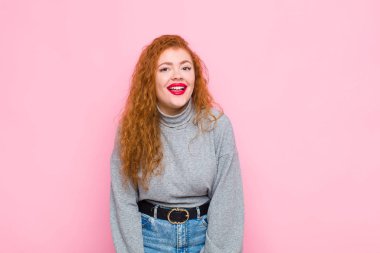 Genç kızıl saçlı bir kadın, büyük, dost canlısı, kaygısız bir gülümseme, pozitif, rahat ve mutlu görünüyor, pembe duvarın karşısında ürperiyor.